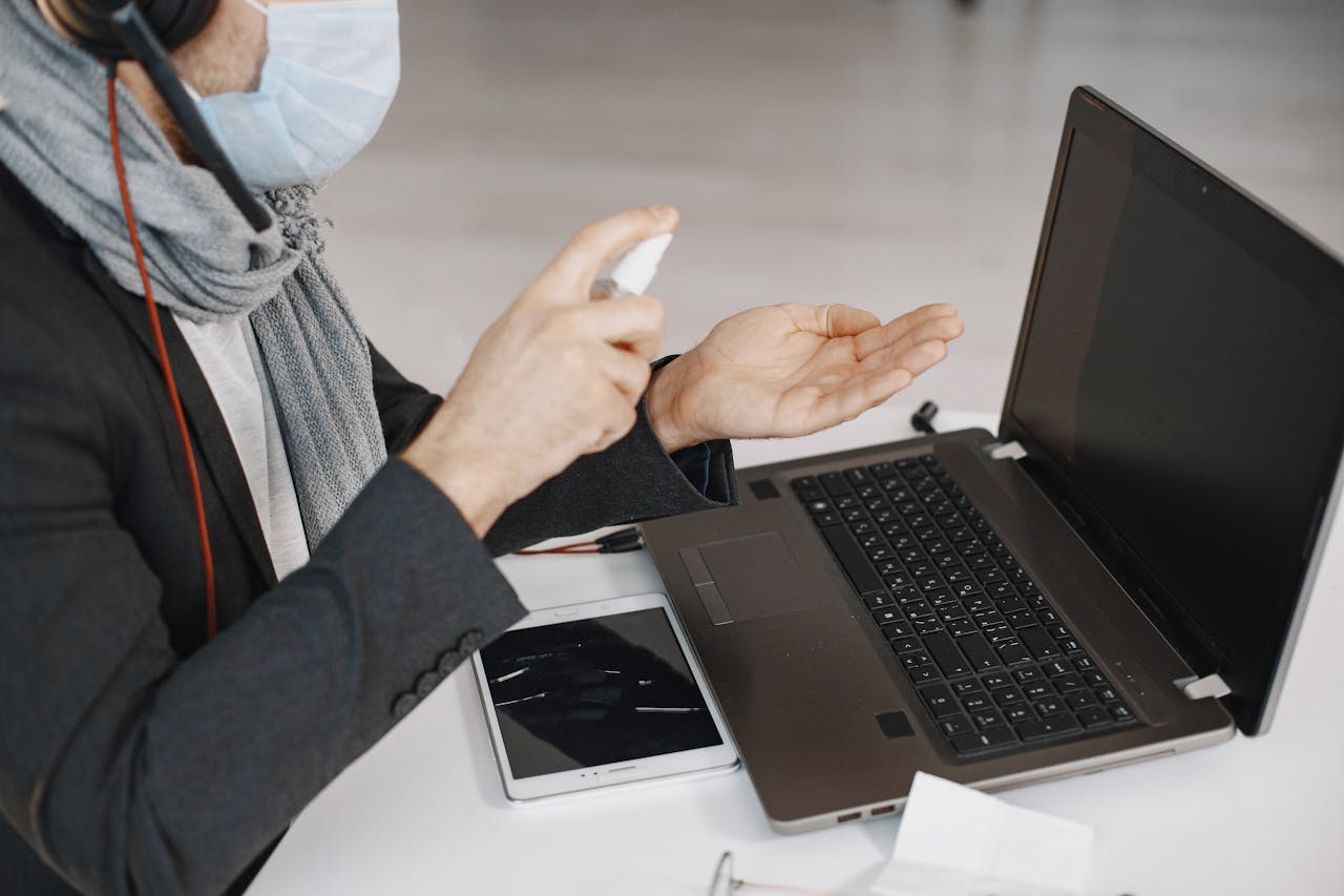 A man wearing a mask sanitizing his hands while working at a laptop indoors.